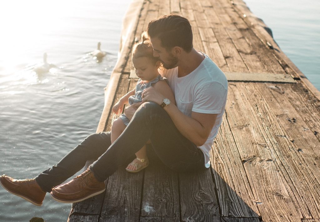 papa portant un t shirt blanc un jean et des mocassins avec sa fille entre les jambes sur un ponton au bord de l'eau