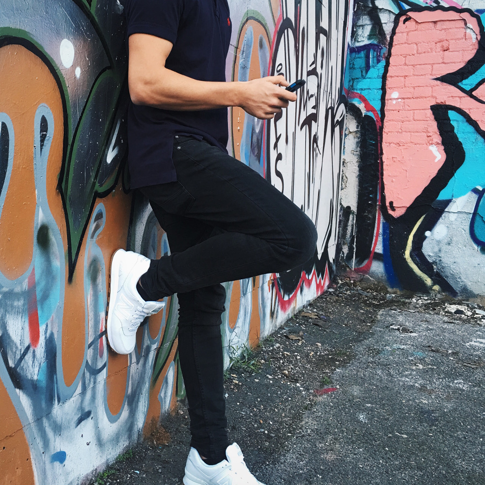 homme adossé à un mur taggé portant un t shirt noir un jean noir et des baskets blanches